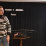 DSC_0336 Prezes Towarzystwa Przyjaciół Ziemi Dobrzańskiej wita naszego gościa. W tle czarna kotara na której widnieje napis „Spotkanie z podróżnika” na stoliku stoją miecze samurajskie, z prawej strony stoi ekran na którym wyświetlane są zdjęcia