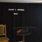 DSC_0339 Mężczyzna w średnim wieku w niebieskiej koszuli i czarnej marynarce stoi przy jasnym krześle. W tle czarna kotara na której widnieje napis „Spotkanie z podróżnika” na stoliku stoją miecze samurajskie, z prawej strony stoi ekran