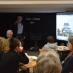 DSC_0345 W bibliotece grupka osób siedzi przy małych stolikach, słuchają mężczyzny który do nich mówi. W tle czarna kotara, stolik na którym stoją miecze samurajskie, z prawej strony stoi ekran na którym wyświetlane są zdjęcia