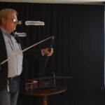 DSC_0350 Mężczyzna w okularach, w niebieskiej koszuli i czarnej marynarce stoi i trzyma w ręku miecz. W tle czarna kotara i ekran.