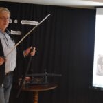 DSC_0352 Stojący mężczyzna w średnim wieku trzyma w ręku miecz samurajski. W tle czarna kotara i ekran.