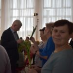 Trzy kobiety dostają po tulipanie od stojącego przed nimi siwiejącego mężczyzny. W tle duże okna z firanami.