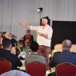 Stoi mężczyzna w białej koszuli i kolorowych spodniach z wyciągniętą prawą ręką. Przed nim siedzi pięć osób na czerwonych krzesłach. W tle orkiestra dęta w czerwono czarnych strojach, czarny tryptyk