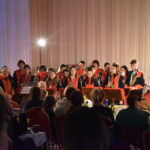 Orkiestra dęta gra na instrumentach przed nimi stoi dyrygent wszyscy są w czarno czerwonych mundurach i siedzą na czerwono złotych krzesłach. W oddali świeci reflektor.