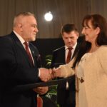 Trzy kobiety dostają po tulipanie od stojącego przed nimi siwiejącego mężczyzny. W tle duże okna z firanami.