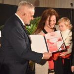 Trzy kobiety dostają po tulipanie od stojącego przed nimi siwiejącego mężczyzny. W tle duże okna z firanami.