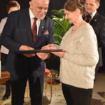 Trzy kobiety dostają po tulipanie od stojącego przed nimi siwiejącego mężczyzny. W tle duże okna z firanami.