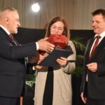 Kobieta w jasnym swetrze otrzymuje z rąk mężczyzny czerwone pudełko, drugi mężczyzna przygląda się na nich