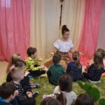 Kobieta w białej bluzce siedzi z rozłożonymi rękami, tłumaczy dzieciom siedzących na zielonym dywanie przed nią. W tle kolorowe kotary.