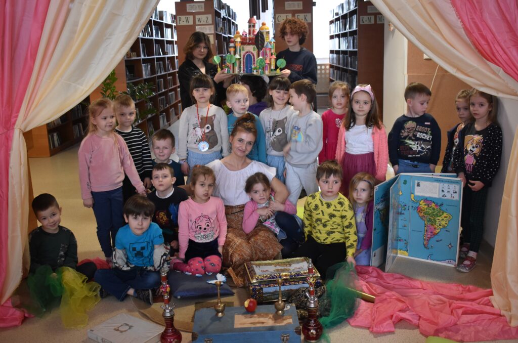 Gróbka przedszkolaków z kobietą w białej bluzce siedzą na dywanie reszta stoi. Przed nimi leżą kuferki czerwone wieże, duży atlas z mapą ameryki południową. Po obu stronach kolorowe kotary