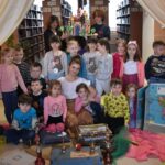 Gróbka przedszkolaków z kobietą w białej bluzce siedzą na dywanie reszta stoi. Przed nimi leżą kuferki czerwone wieże, duży atlas z mapą ameryki południową. Po obu stronach kolorowe kotary