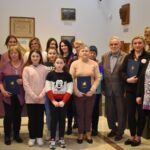 Dwaj mężczyźni w garniturach, w środku kobieta w kremowej sukience.