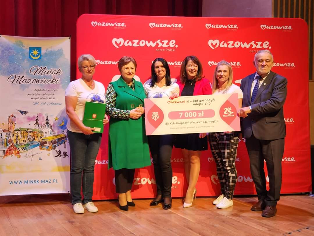 kolka kobiet stojących obok sienie, w tle czerwona sianak, usmiech, obok mezczyzna w garniturze