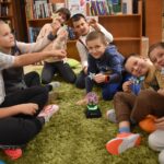 Na zielonym dywanie stoi kula a dzieci siedzące w kole dotykają ją rękoma