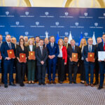 Obraz przedstawia grupę osób stojących w dwóch rzędach, ubranych elegancko, większość mężczyzn w garniturach, a kobiety w sukienkach lub eleganckich garniturach. Na pierwszym planie sześć osób trzyma certyfikaty lub podobne dokumenty w brązowych okładkach. W tle stoi drugi rząd ludzi, również z dokumentami w rękach. Wszyscy patrzą w stronę kamery z uśmiechem i wyglądają na zadowolonych. Tło stanowi wnętrze o eleganckim charakterze, prawdopodobnie sala konferencyjna lub podobne pomieszczenie. Na ścianie w tle widnieje kilka flag, co może sugerować, że jest to wydarzenie o charakterze międzynarodowym lub rządowym. Pośrodku, na ścianie, znajduje się duże logo lub herb, co może wskazywać na konkretną instytucję lub organizację. Na podłodze jest wykładzina, a całe pomieszczenie jest dobrze oświetlone. Wydarzenie to wydaje się być ważną ceremonią lub uroczystością, być może wręczeniem nagród, certyfikatów lub uznania dla grupy ludzi. Atmosfera jest formalna i celebracyjna.