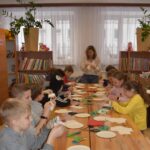 92 Po obu stronach drewnianego stołu siedzą dziewczynki i chłopcy, na końcu stołu stoi kobieta i pokazuje im jak robić kwiaty z bibuły. W tle Okno i regały z książkami