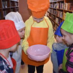 187 Pięć dziewczynek stoi w kolorowych czepkach kucharskich i zagląda do różowej miski. W tle regały z książkami