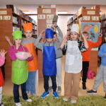 195 Czterech chłopców i cztery dziewczynki stoi w kolorowych fartuszkach trzymają w rękach miskę, deskę i tarkę. W tle regały biblioteczne z książkami