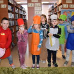 198 W bibliotece dzieci szykują się do zajęć kulinarnych ubrane w fartuszki i czepki kucharskie z miską, tarką i deskami do krojenia. W tle regały z książkami