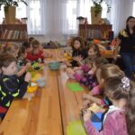 212 Dzieci siedzą przy stole przed nimi deski do krojenia, miski. W tle regały z książkami