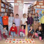 221 Grupa dzieci pozuje do zdjęcia pierwszy rząd kuca drugi stoi, przed nimi leżą laurki, które zrobili dla swoich Babć. W tle regały z książkami.