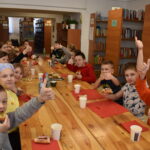 246 Dzieci siedzą przy stole i jedzą zrobione przez siebie kanapki. W tle regały z książkami