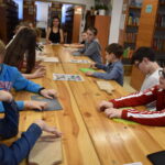 262 Przy drewnianym stole siedzą dzieci, na końcu siedzi kobieta. Przed nimi leżą deski do krojenia, w tle regały z książkami.