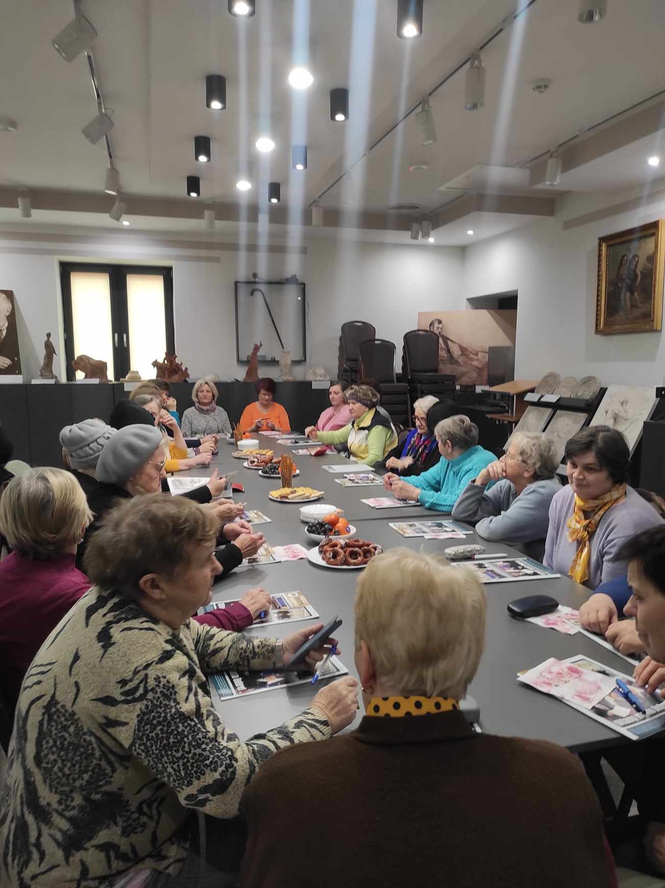 Na obrazku grupy osób siedzących wokół stołu. Wyglądają na to, że są zaangażowane w danie lub posiłek, ponieważ niektóre z nich mają przed sobą talerz lub dokumenty.