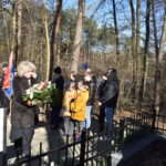 Na zdjęciu jest grupa osób stojących przed ogrodzeniem. Wyglądają na skoncentrowanych, zwracając swoją uwagę ku temu, co kryje się za płotem.