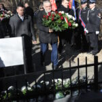 Na obrazku widać grupę osób stojących przed ogrodzeniem. Wyglądają na podejrzane lub skupione, podejrzanec, że może wykryć coś poza klastką.