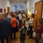 Na udostępnienie jest grupa ludzi, która stoi w przerwie i szerokim korytarzu. Osoby te mają sobie różne ubrania i wyglądają na to, że są w trakcie rozmów lub oczekiwania na coś.