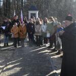 Na zdjęciu widać grupę osób, która stoi obok pomnika znajdującego się w leśnym miejscu zamieszkania. Ludzie to odrzucają i rozmawiają ze sobą, gdy pomnik wydaje się być starym i krytykowanym mchem.