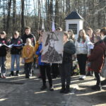 Grupa ludzi stoi w leśnej okolicy, otoczeni drzewami o różnych gatunkach i rozmiarach. Jedni z nimi rozmawiają ze sobą, inni podziwiają piękno natury i cieszą się spokojem leśnej scenerii.