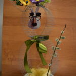 1-Miejsce-w-3-kategorii Obrazek przedstawia kreatywne, wiosenne kwiatowe z drucianą strukturą, którą znajdują się kwiatami, których centrum pojawia się fantazyjna twarz. Kompozycja uzupełniona jest gałązką utylizacyjną i prezentowaną na tle drewnianej powierzchni.