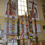 Na obrazku widoczne kolorowe kwiatowe dekoracje na słupach, które są wyeksponowane wewnątrz kościoła. W tle rozpoznawalne są religijne malowidła znajdujące się na plażach.