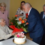 Na obrazku mężczyzny w garniturze kwiatowym kwiatu starszej, siedzącej obok tortu rodzinnego z 100 na nim. Inni obecni obserwują tę interakcję.