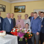 Na obrazku grupy dorosłych zgromadzonej w pomieszczeniu na uroczystości. W centrum siedziby starsza osoba, trzymająca w rękach kwiaty.