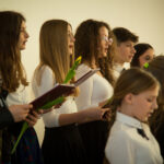 Na obrazku grupę dziewcząt, które śpiewają w chórze. Ubiorone są jednymi w jednolitych strojach, które są przeznaczone na dyrygencie, które prowadzą ich wykonanie.