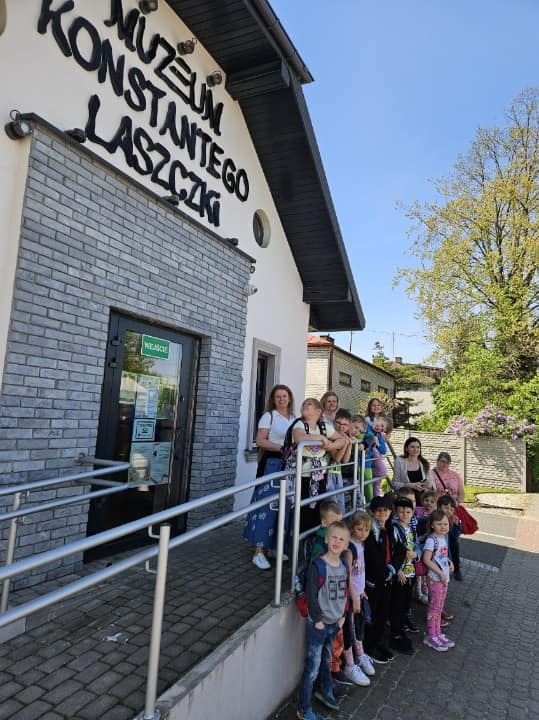 Nagranie wideo jest grupą dzieci i dorosłych, które stoi na zewnątrz wejścia do muzeum Konstantego Laszczki w słoneczny dzień. Wszyscy szczególni są narażeni na widoczność, a budynek muzeum jest dobrze oświetlony dzięki światłu słonecznemu.