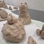 Na obrazku są gliniane figurki zwierząt, które występują na białym tle. Zwróć uwagę na siedzącą figurę kota, a zaraz przed nią pojawi się mały, zaokrąglony model.