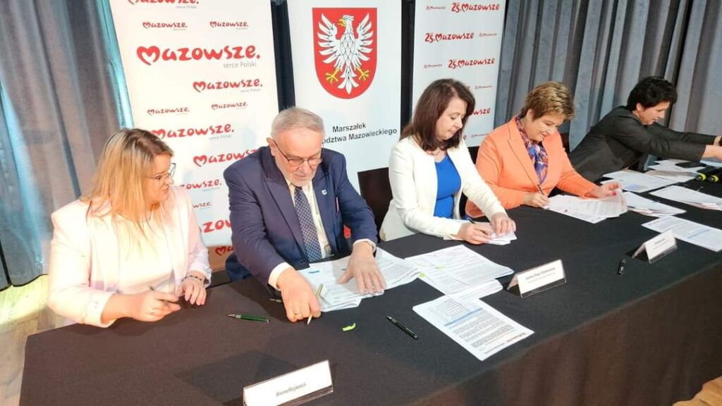 Na ekranie następuje odbiór dokumentów podczas formalnych wydarzeń. W tle widoczne są banery z logiem Mazowsza.