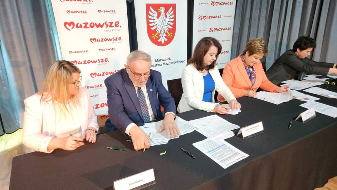 Na ekranie następuje odbiór dokumentów podczas formalnych wydarzeń. W tle widoczne są banery z logiem Mazowsza.