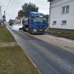 Na obraz widoczny jest samochód do czyszczenia ulicy, który rozpyla wodę na drogę. W tle można zrelaksować się domy oraz dodatkowe dodatkowe, a wszystko, co dzieje się podczas pochmurnego dnia.