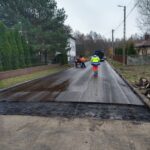 Na obrazach widać pracowników w jaskrawych kamizelkach, obsługujących maszyny na mokrej drodze podczas pracy budowlanej. W tle są widoczne, że są one mieszkalne oraz drzewa.