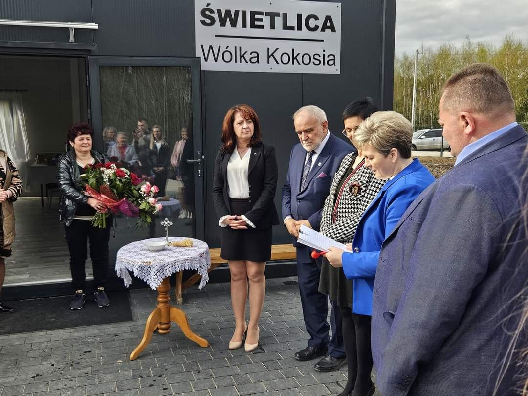 Na zdjęciu grupa osób, która stoi na zewnątrz budynku podczas formalnej ceremonii, niektóre z nich trzymają papiery w rękach. W pierwszym rzędzie znajduje się składnik, na którym znajduje się składnik ziołowy.