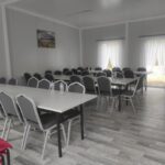 Zdjecie-2 Obrazek przedstawia pustą salę konferencyjną z szarymi stołami i krzesłami na drewnianym wyposażeniu. Położenie jest jasne dzięki naturalnemu światłu wpadającemu przez zasłonięte okna.