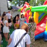 1716985030928 Na obrazku grupy dorosłych i dzieci zgromadzonych wokół dmuchanego zamku do skakania na świeżym powietrzu. Część osób czekających w kolejce, podczas gdy inni obserwują z boku; w tle widać drzewa i budynek.