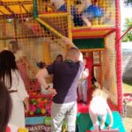 1716985030934 Dzieci bawią się na kolorowej konstrukcji placu zabaw z siatkami i miękkimi powierzchniami, gdy są dostępne w zasięgu sieci. Dziewczynka w niebieskiej sukience wspina się, a inne dziecko stoi na pochylni.