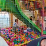 1716985030940 Na obrazku dzieci bawią się w gry z rzeczywistymi urządzeniami do gry w piłkę nożną, a nad nimi znajdują się zielona zjeżdżalnia. widoczna jest otoczona siatkowa konstrukcja wewnątrz sali zabaw.