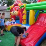 1716985030954 Na obrazku dzieci bawią się na dmuchanej zjeżdżalni ozdobionej motywami lwów, przy użyciu dorosłych. Kobieta pomaga jednemu z dzieci wspinać się na zjeżdżalnię, podczas gdy reszta czeka w pobliżu; całość odbywa się w plenerze.